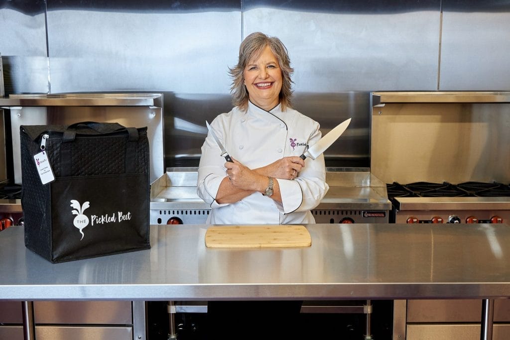 The Pickled Beet Founder-CEO Chef Liz standing in its gluten-free commercial kitchen, wearing white chef coat and holding chef knives